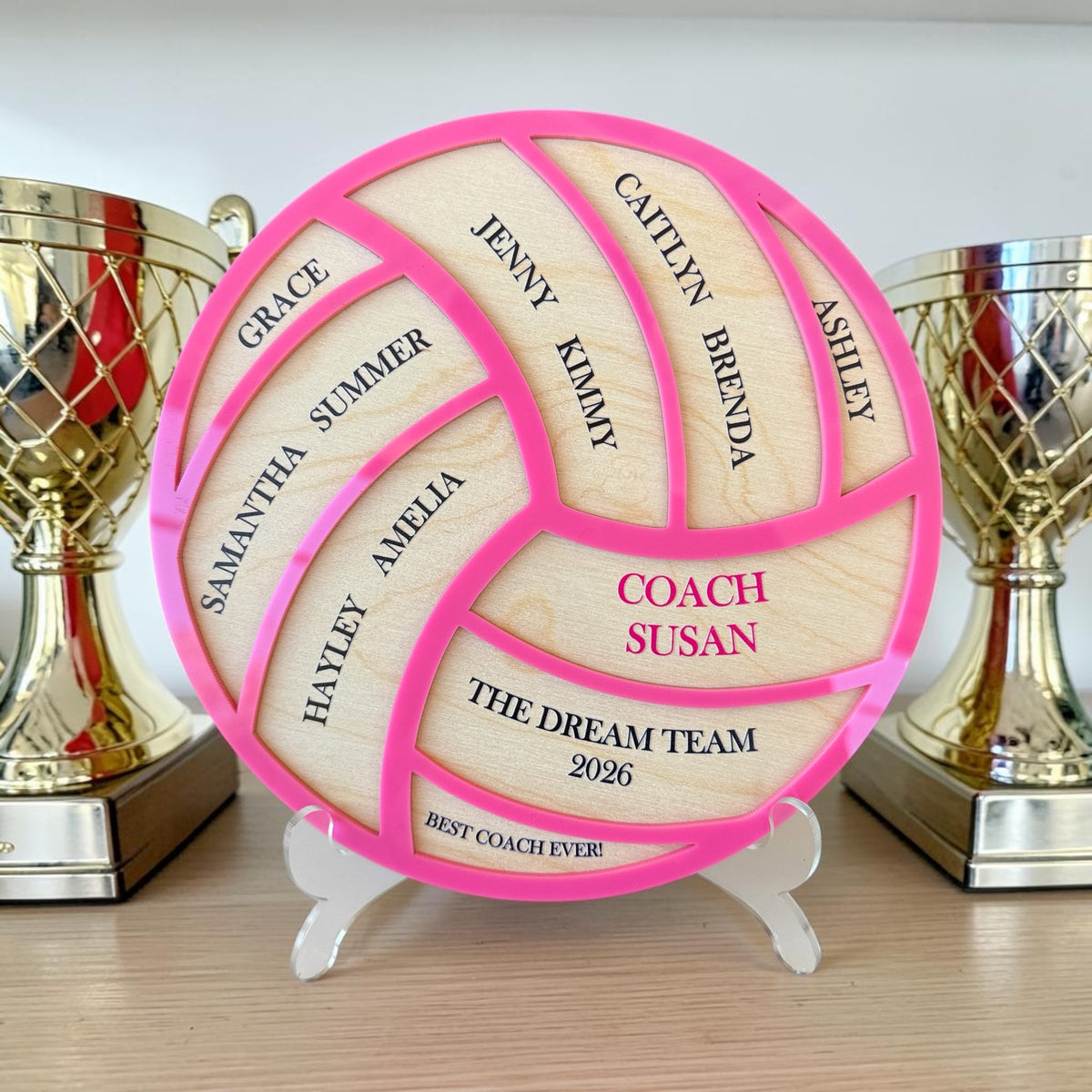 A custom wooden volleyball-shaped plaque with bright pink borders, displayed on a small white stand. The natural wood sections are engraved with names and text including 'COACH SUSAN', 'THE DREAM TEAM 2026', and 'BEST COACH EVER'.