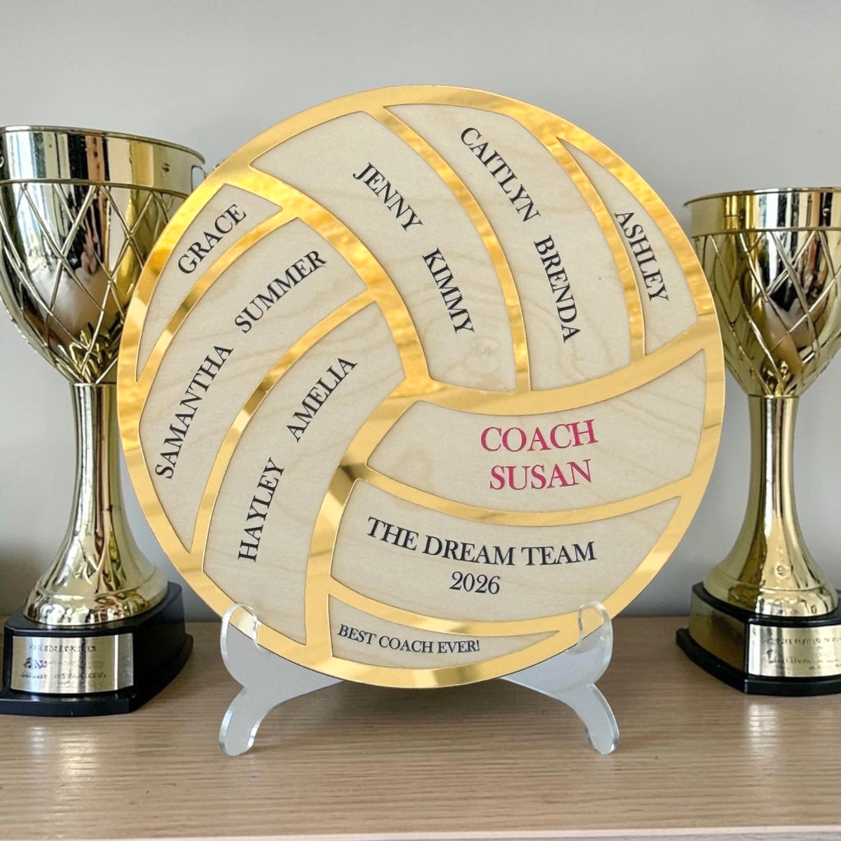 A round wooden plaque shaped like a volleyball, personalized with names and the text 'COACH SUSAN' and 'THE DREAM TEAM 2026', displayed on a small white stand between two gold trophy cups on a shelf.