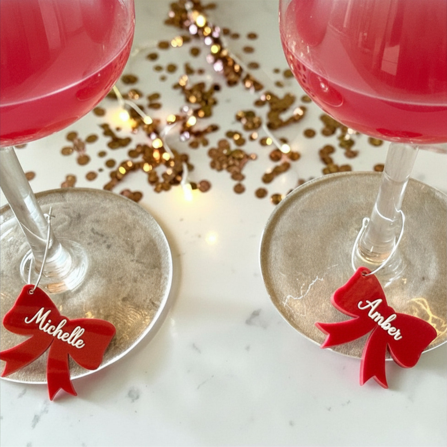 Two stemmed wine glasses filled with pink drink, placed on a white marble surface with scattered gold confetti and fairy lights. Each glass has a small red bow-shaped tag attached near the base, one labeled "Michelle" and the other "Amber.
