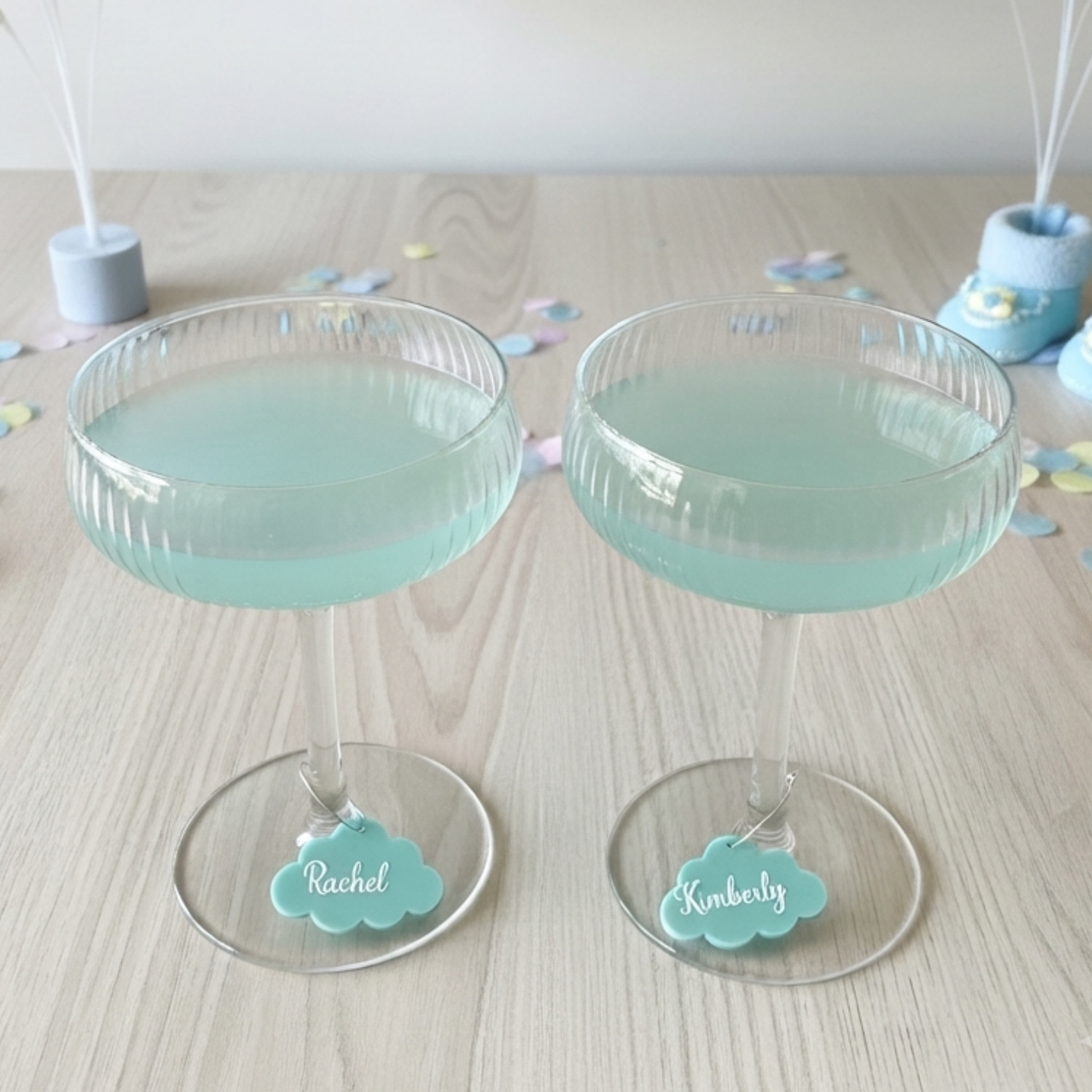 Two clear stemmed coupe glasses filled with pale aqua liquid, placed on a light wood tabletop. Each glass has a small cloud-shaped tag attached near the base, one labeled Rachel and the other Kimberly.