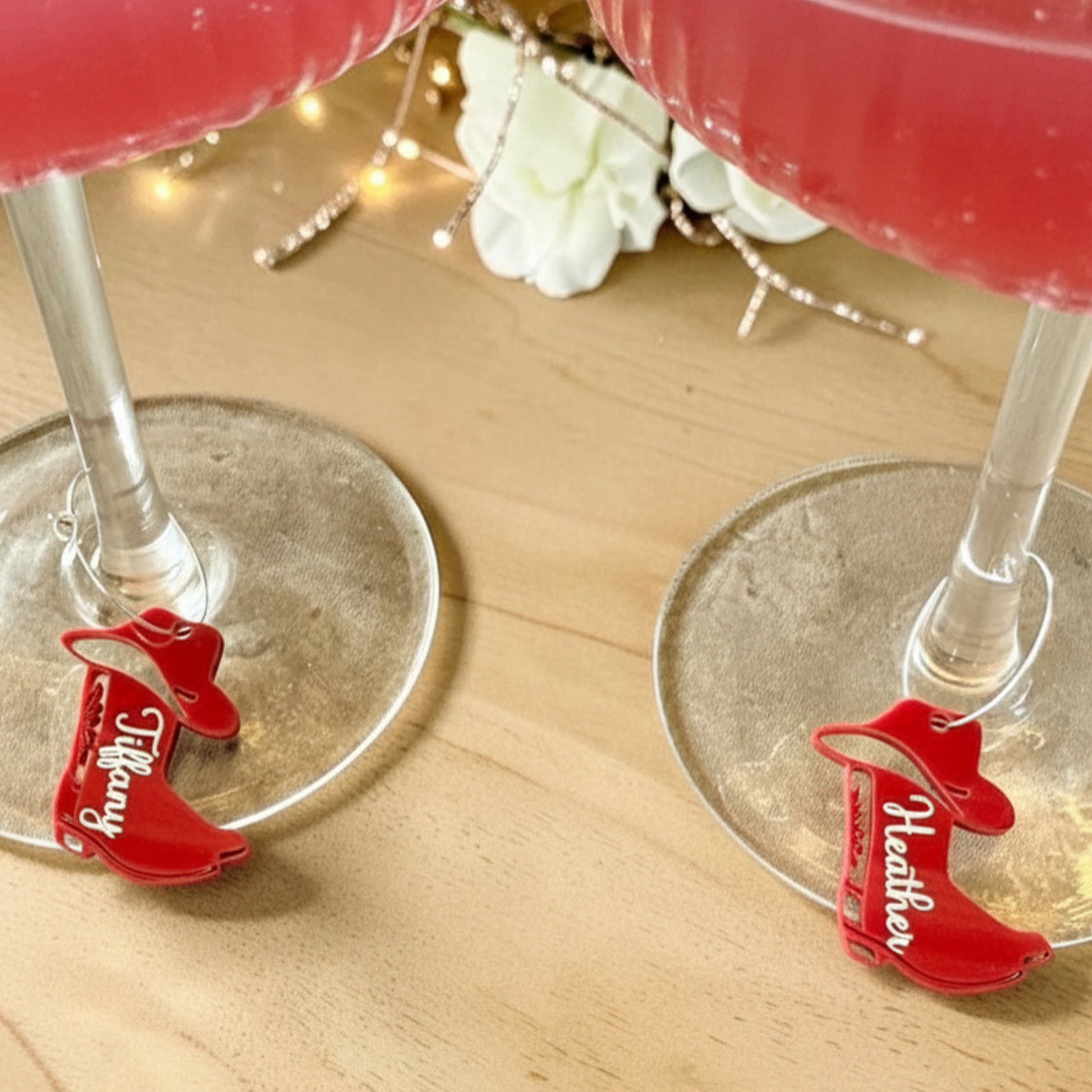 Two red high-heeled shoe-shaped cocktail glasses placed on a light wood surface, each holding a pink drink. The left shoe-shaped glass has 'Tiffany' in white script, and the right one has 'Heather' in white script.