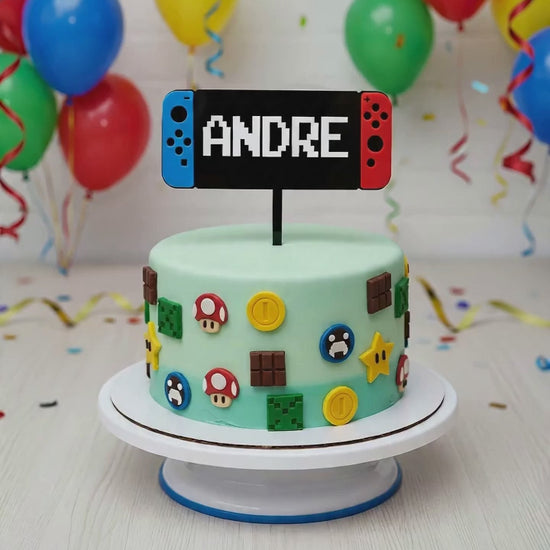 A personalised laser-cut acrylic gaming cake topper, shaped like a red and blue handheld video game console with the custom name "ANDRE" in a white pixelated font. The 14-15cm wide topper is displayed on a light green birthday cake decorated with Mario and Minecraft fondant shapes.