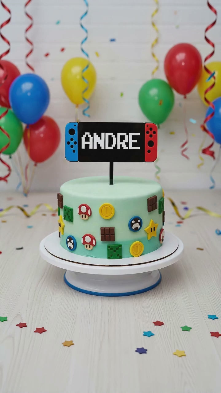 A personalised laser-cut acrylic gaming cake topper, shaped like a red and blue handheld video game console with the custom name "ANDRE" in a white pixelated font. The 14-15cm wide topper is displayed on a light green birthday cake decorated with Mario and Minecraft fondant shapes.
